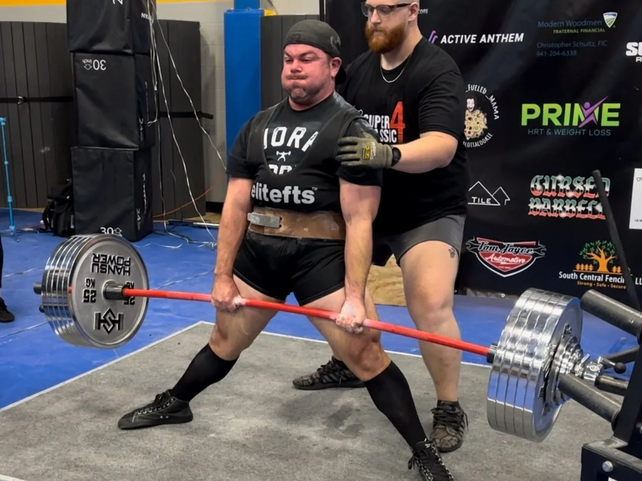 A man in powerlifting gear is performing a deadlift with a loaded barbell, while another man in black shorts and t-shirt stands behind him, spotting and supporting during the lift in a gym setting.