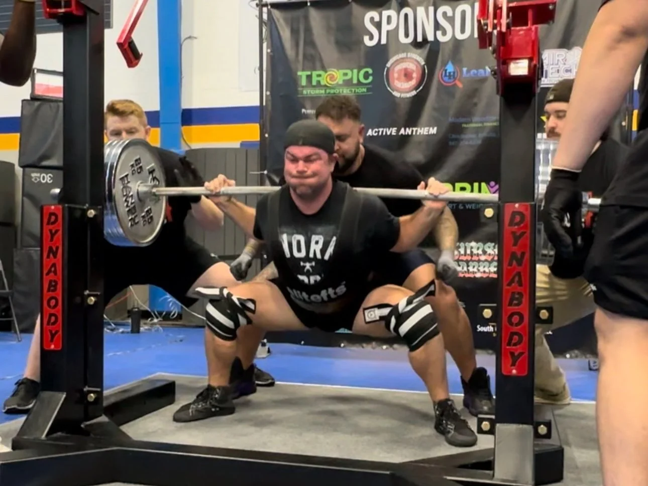 A man performing a squat exercise with a barbell at a powerlifting event, surrounded by spotters and gym equipment.
