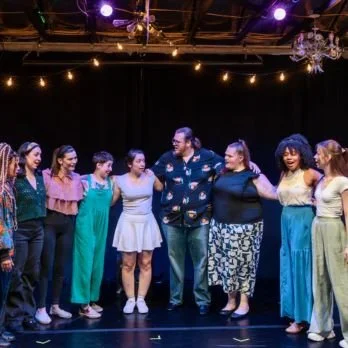 A group of seven women and one man stand on a stage, holding hands and smiling, under string lights.
