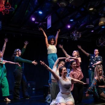 Group of diverse women performing a dance on a stage with dark background and colorful lights.