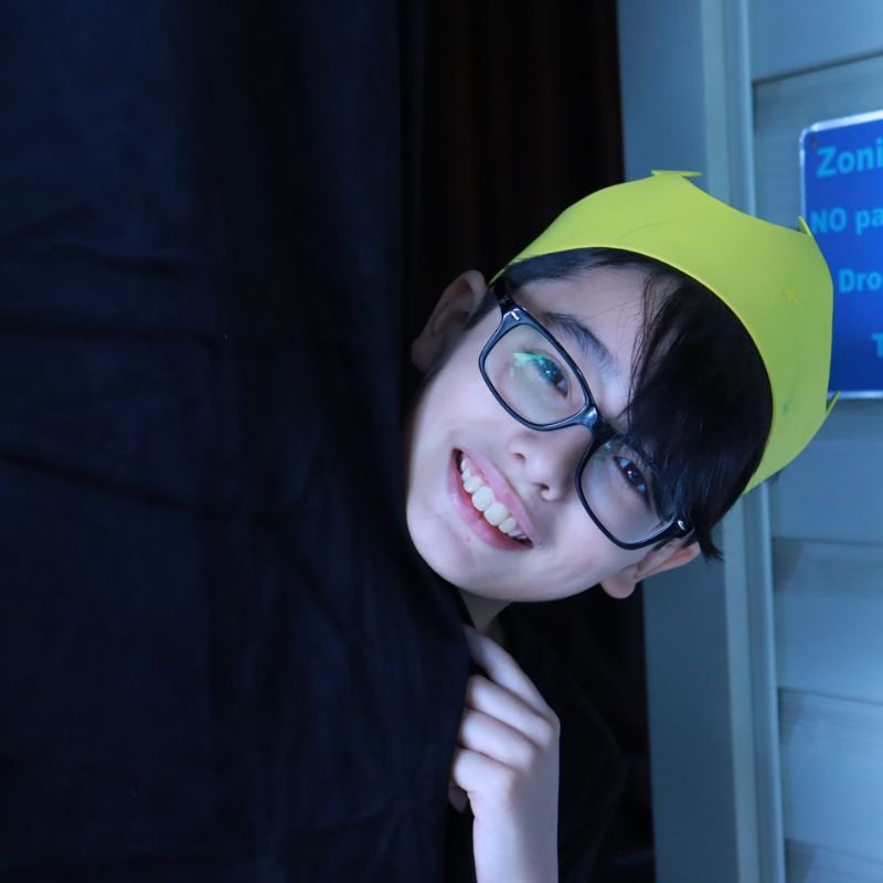A young person wearing glasses and a yellow paper crown, smiling and leaning against a dark surface, with a blue zone sign visible in the background.