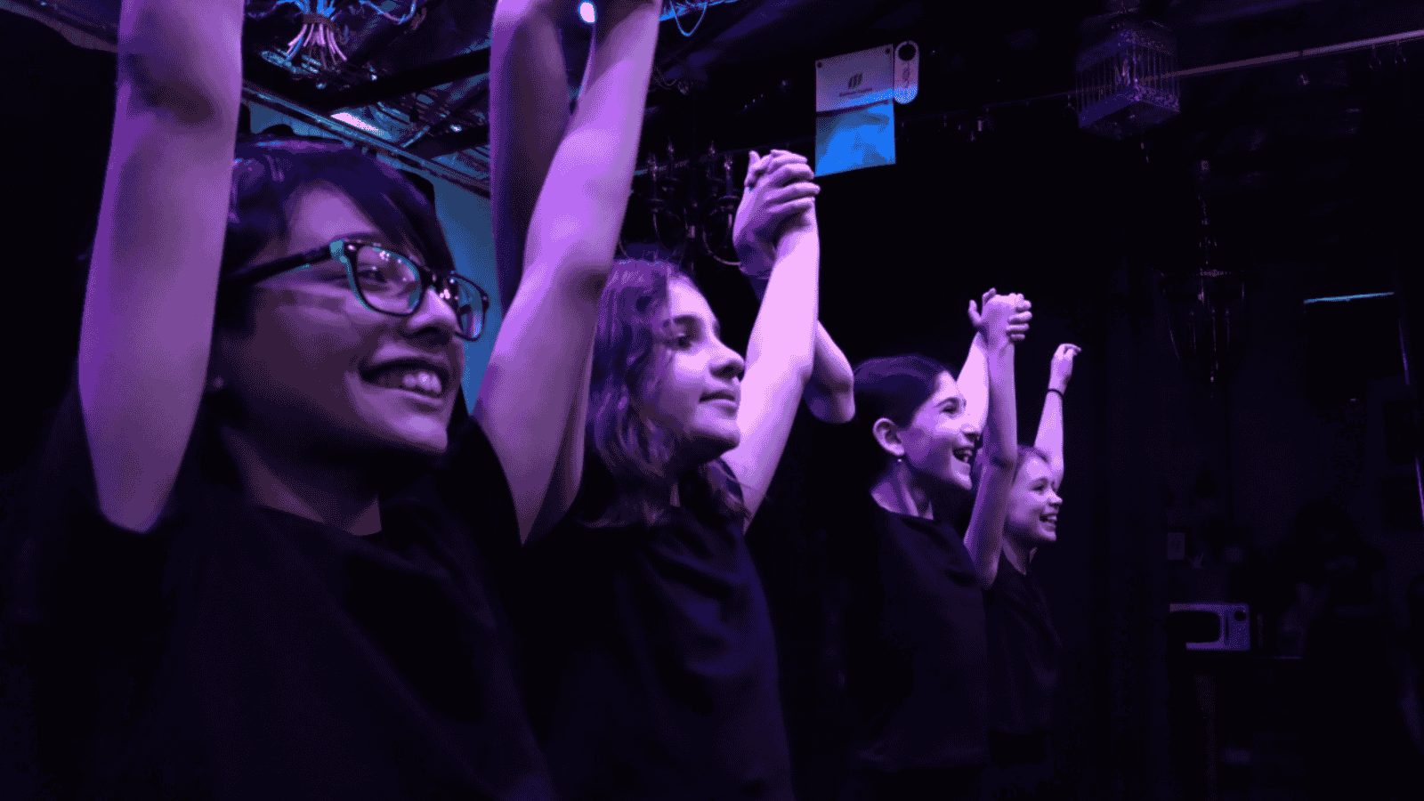 Four young women on stage with their hands raised, smiling, under purple stage lighting, at a live event or performance.