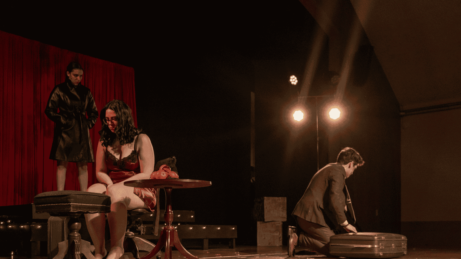 Three performers on stage during a theater performance; one standing near a red curtain, one sitting with glasses and a red dress, and a third kneeling packing a suitcase, illuminated by stage lights.