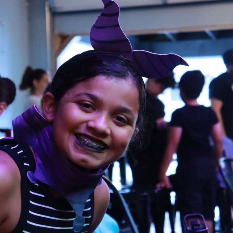 Child wearing a witch's hat with purple and black stripes, smiling with braces, indoors with people in the background.