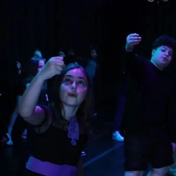 Children standing on a stage, with one girl looking up and holding her hand near her head, and a boy raising his arm.