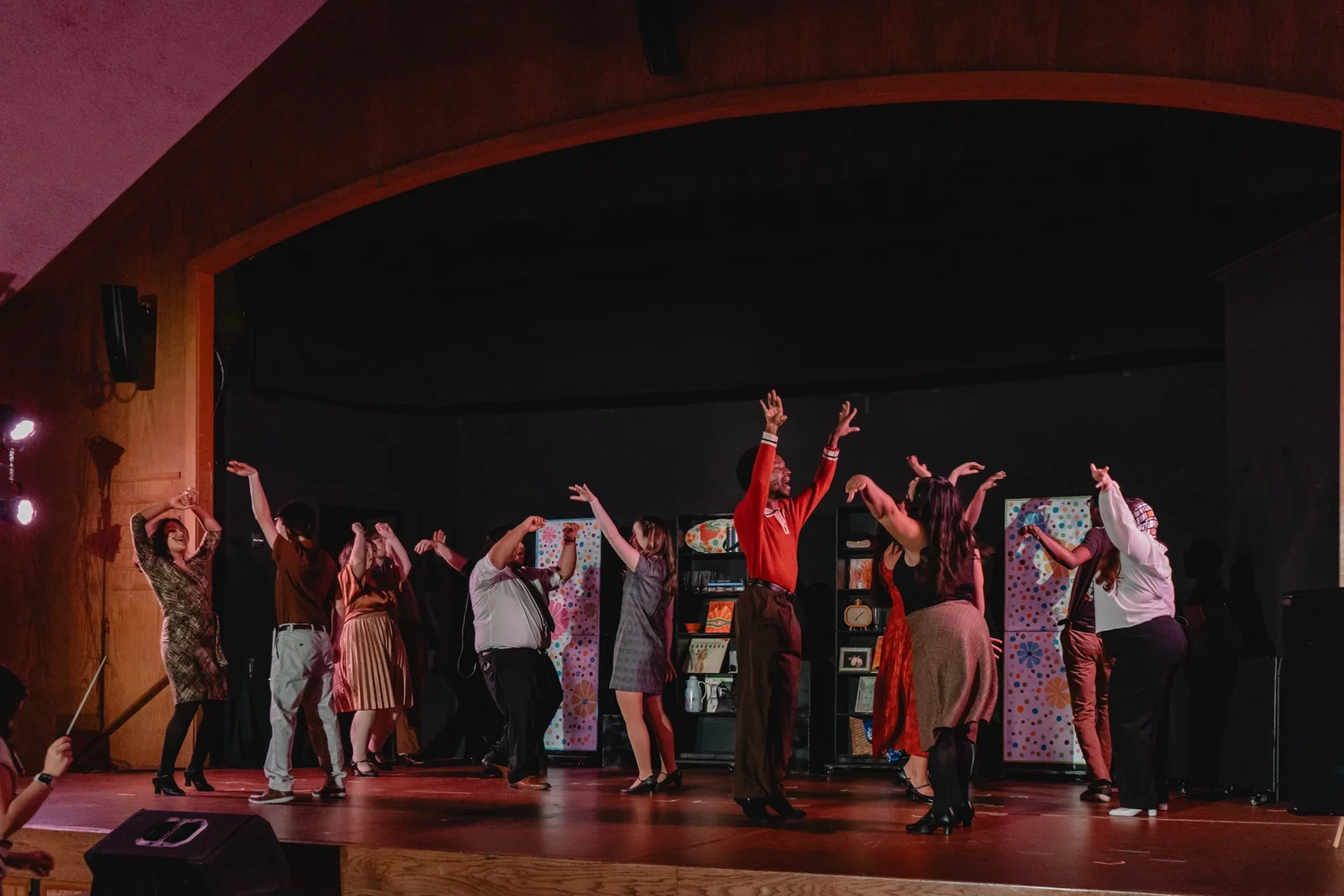 The cast of Merrily We Roll Along dancing with their arms raised across a wide proscenium stage