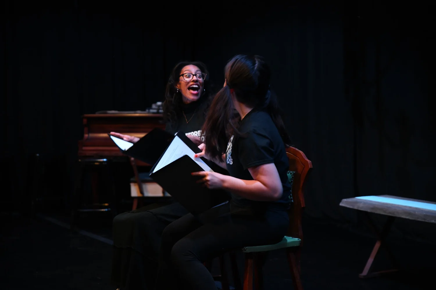 Two performers reading from black script binders beside a piano, one of them laughing