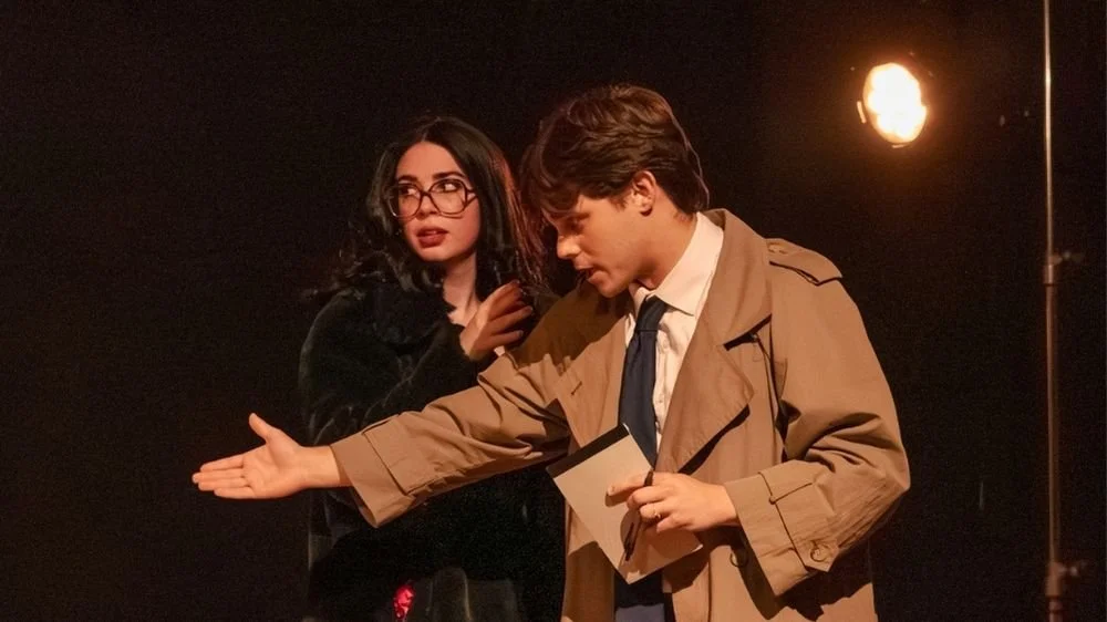 A woman with glasses and dark hair wearing a black outfit and a man with brown hair wearing a tan trench coat and tie on a stage, with stage lights in the background.