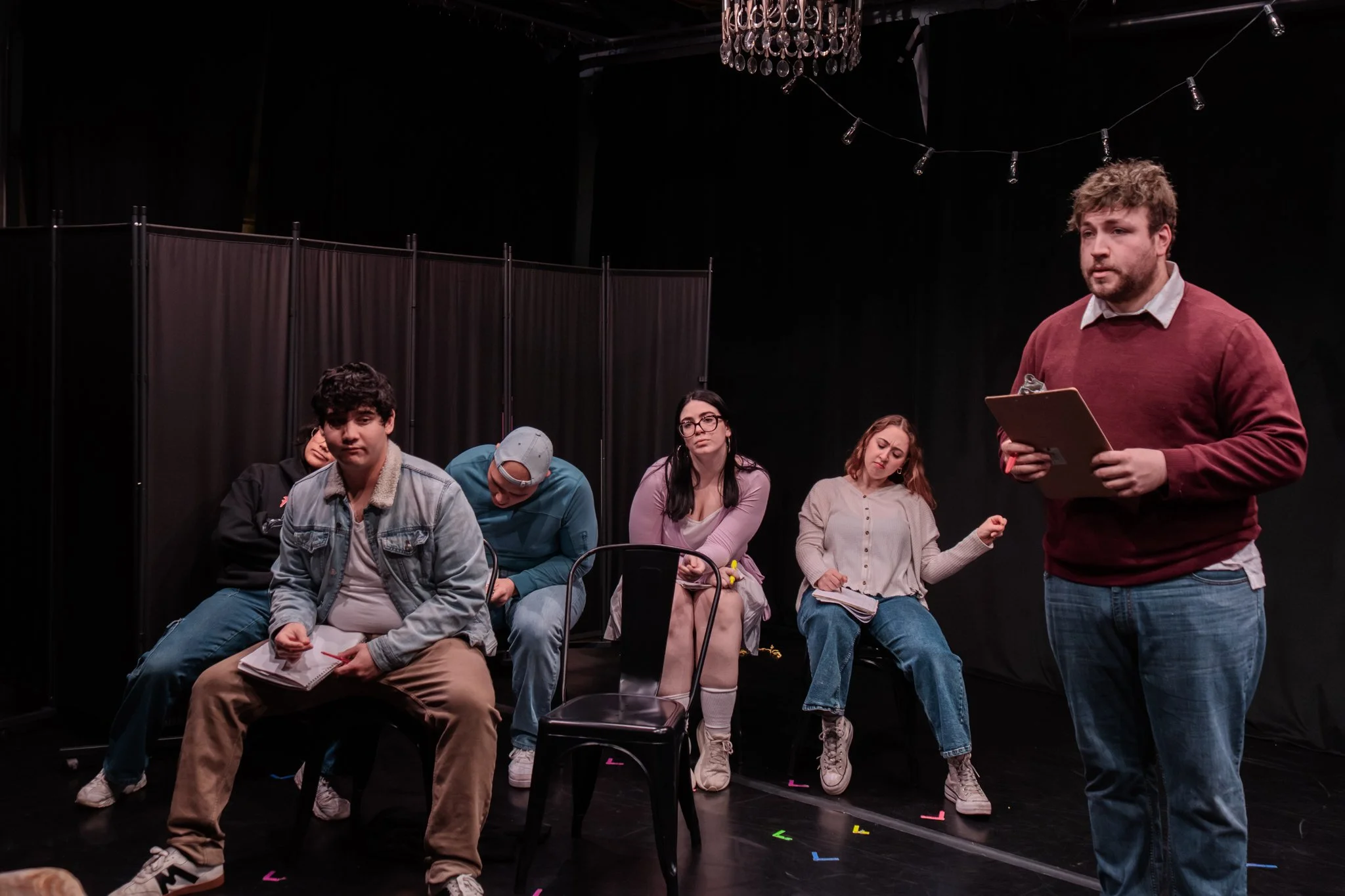 A group of six young people seated on stage, with a man standing at the front holding a clipboard, likely portraying a director or instructor in a theater rehearsal or class.