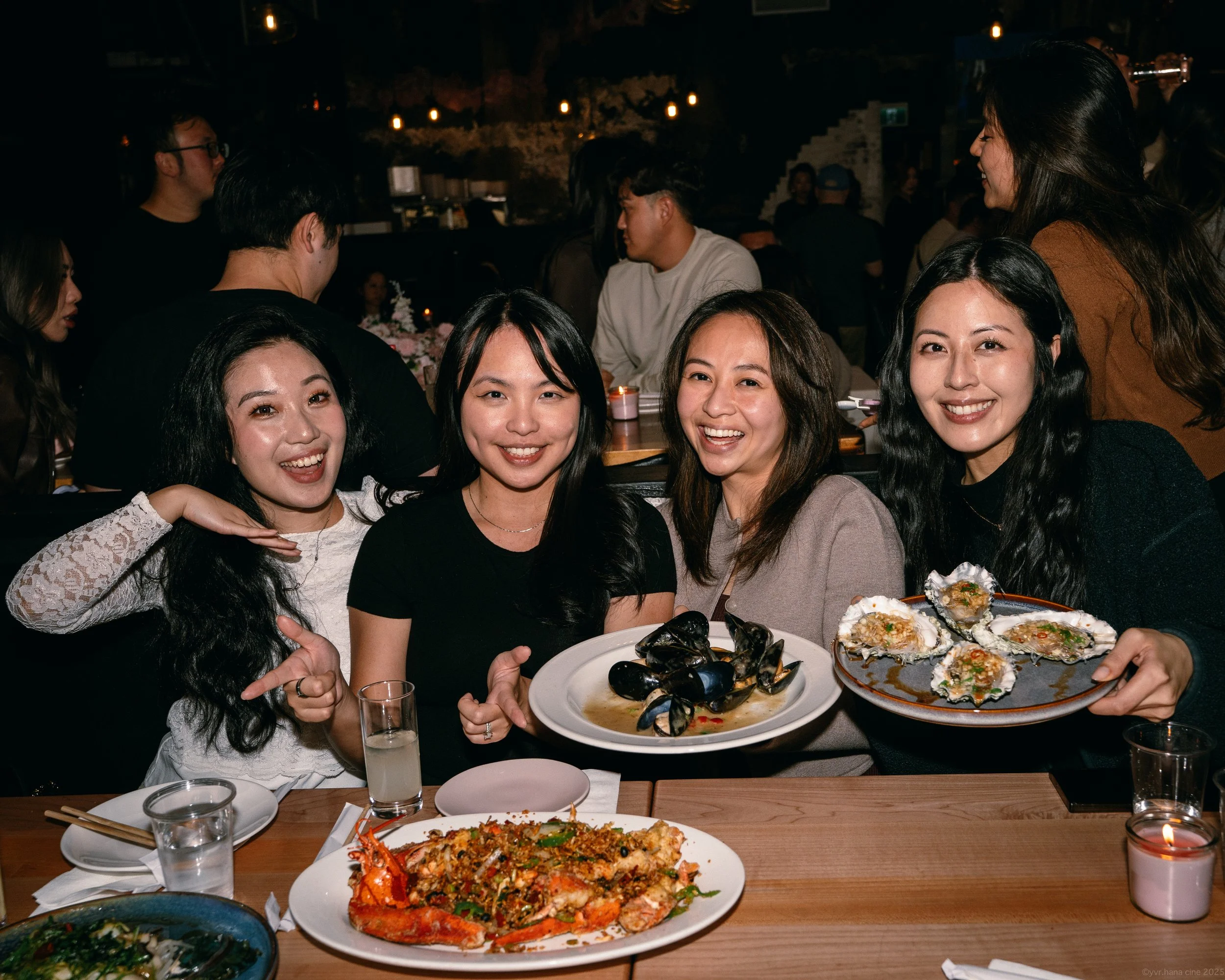 Four women smiling at a dinner table with plates of seafood, including oysters and a lobster dish, in a lively restaurant setting.