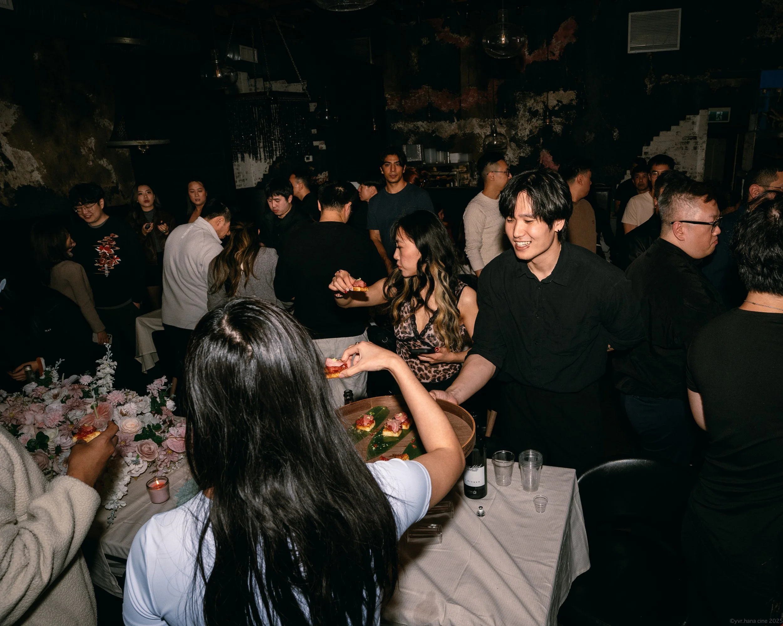 People at a party exchanging food and drinks in a dimly lit indoor space with a rustic, textured wall.