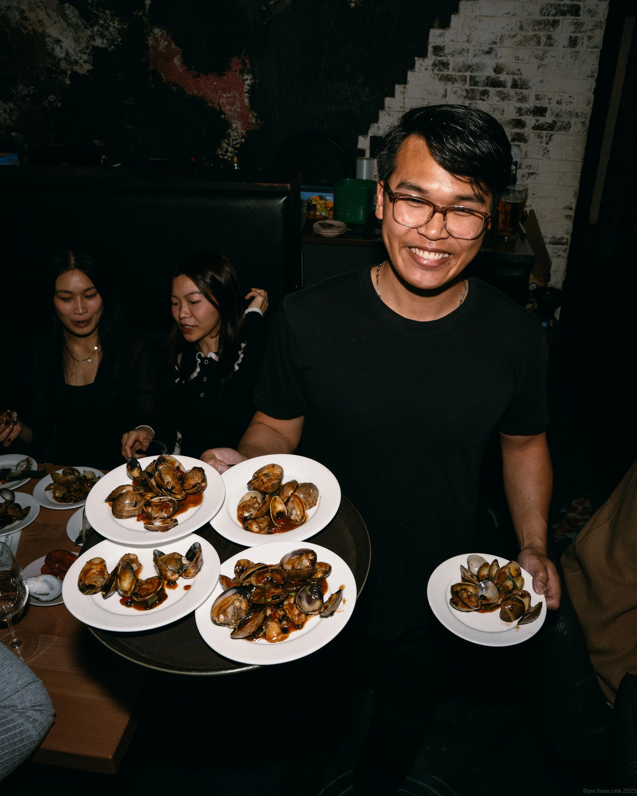 A man smiling and holding a tray of cooked clams at a restaurant, with two women sitting at the table behind him.