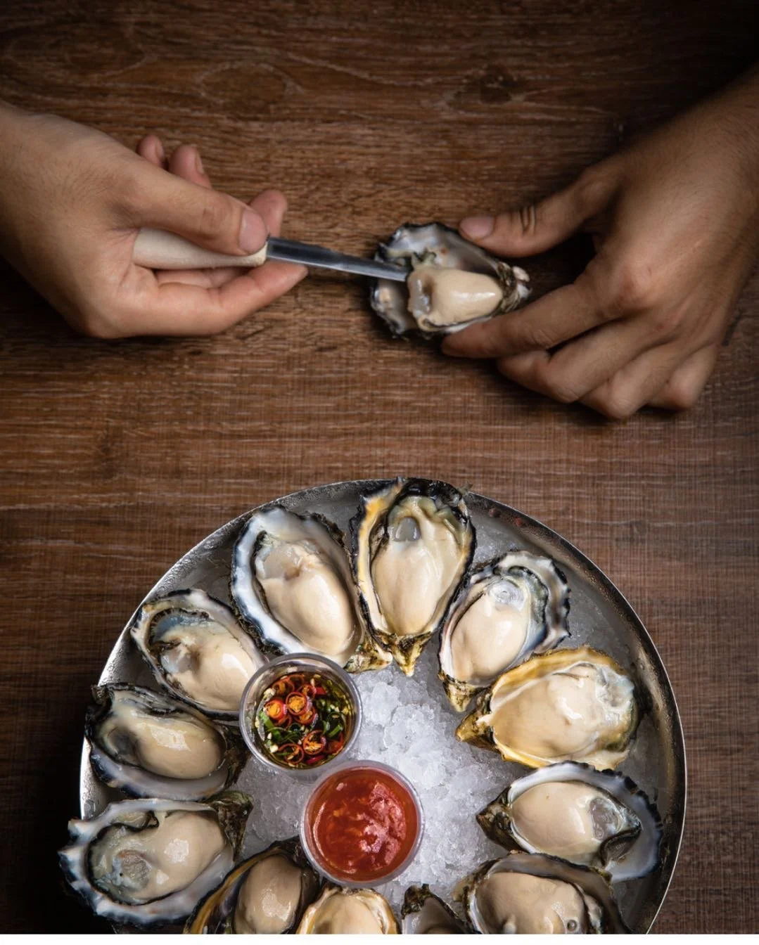 Hands shucking fresh oysters on a wooden table with two dipping sauces on a tray of ice.