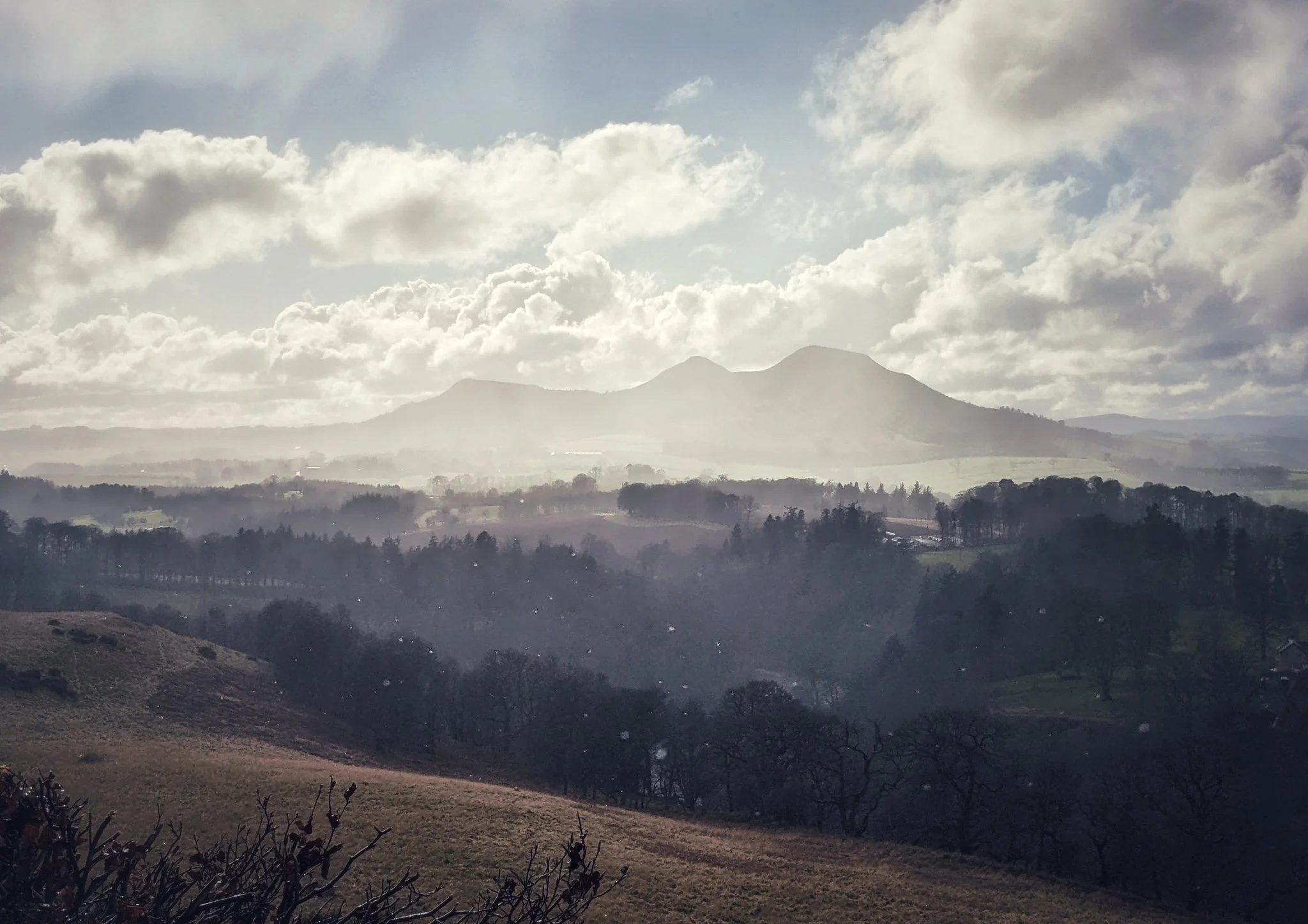 The three peaks of the Eildon Hills rise up from fertile fields and forests, almost glowing due to recently fallen rain and the sun emerging from the clouds behind them.