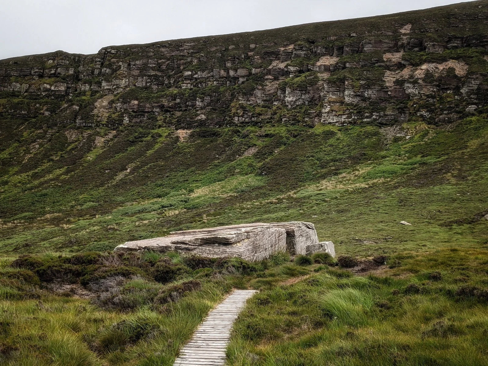 A wooden gangway leads to a massive slab of stone with a 'door' in its side, backed by a sheer wall of cliffs and green-brown grass. Dwarfie Stane, Hoy, Orkney.
