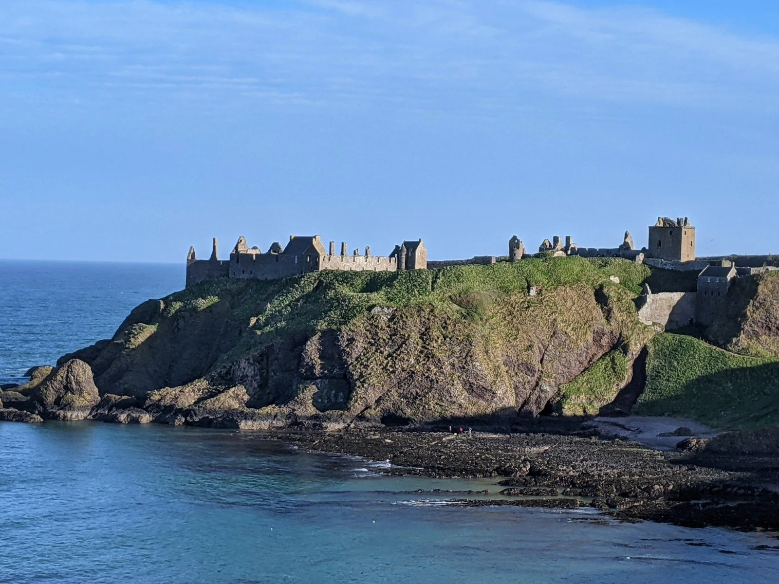 View across a bay to a massive castle atop a clifftop promontory, with sheer drops on all sides. Dunnottar Castle, Stonehaven.