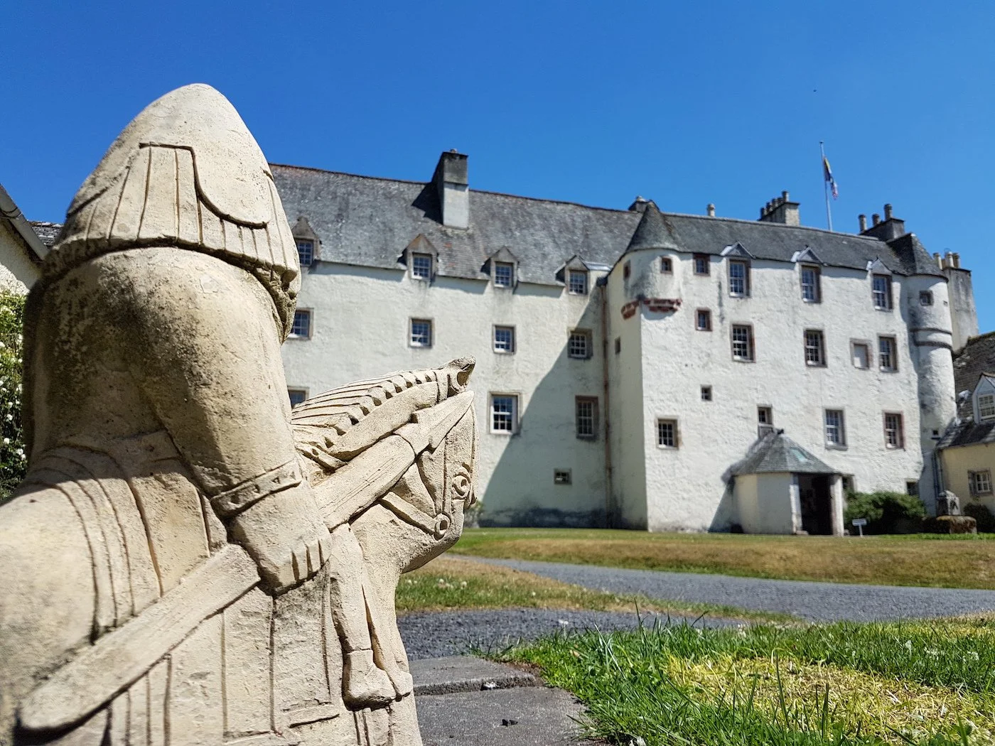 A garden figure based on the Lewis Chessmen, atop a horse and clutching a spear, looks away and towards a white-harled, 3-storey castle. Traquair House, Scottish Borders.