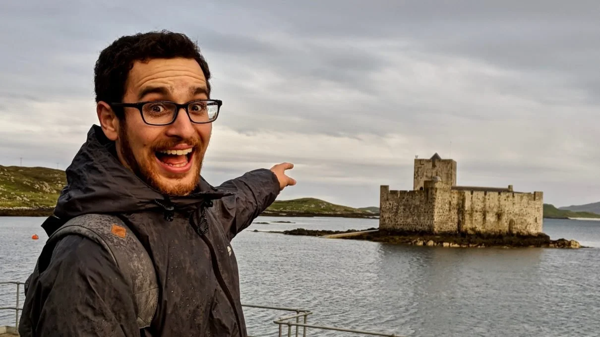 David looking at the camera, wearing a black raincoat, pointing excitedly at a castle lapped by waves in a bay. Kisimul Castle, Barra.