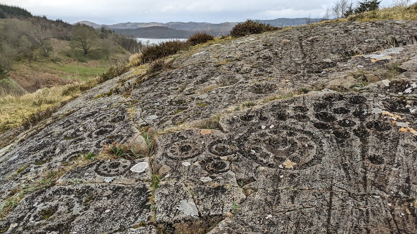 Low angle view across a rocky outcrop at Ormaig, Argyll, covered in abstract rock art designs, including saucer-like hoops, rings, and cup marks. Green hills and a waterway peek out in the background.