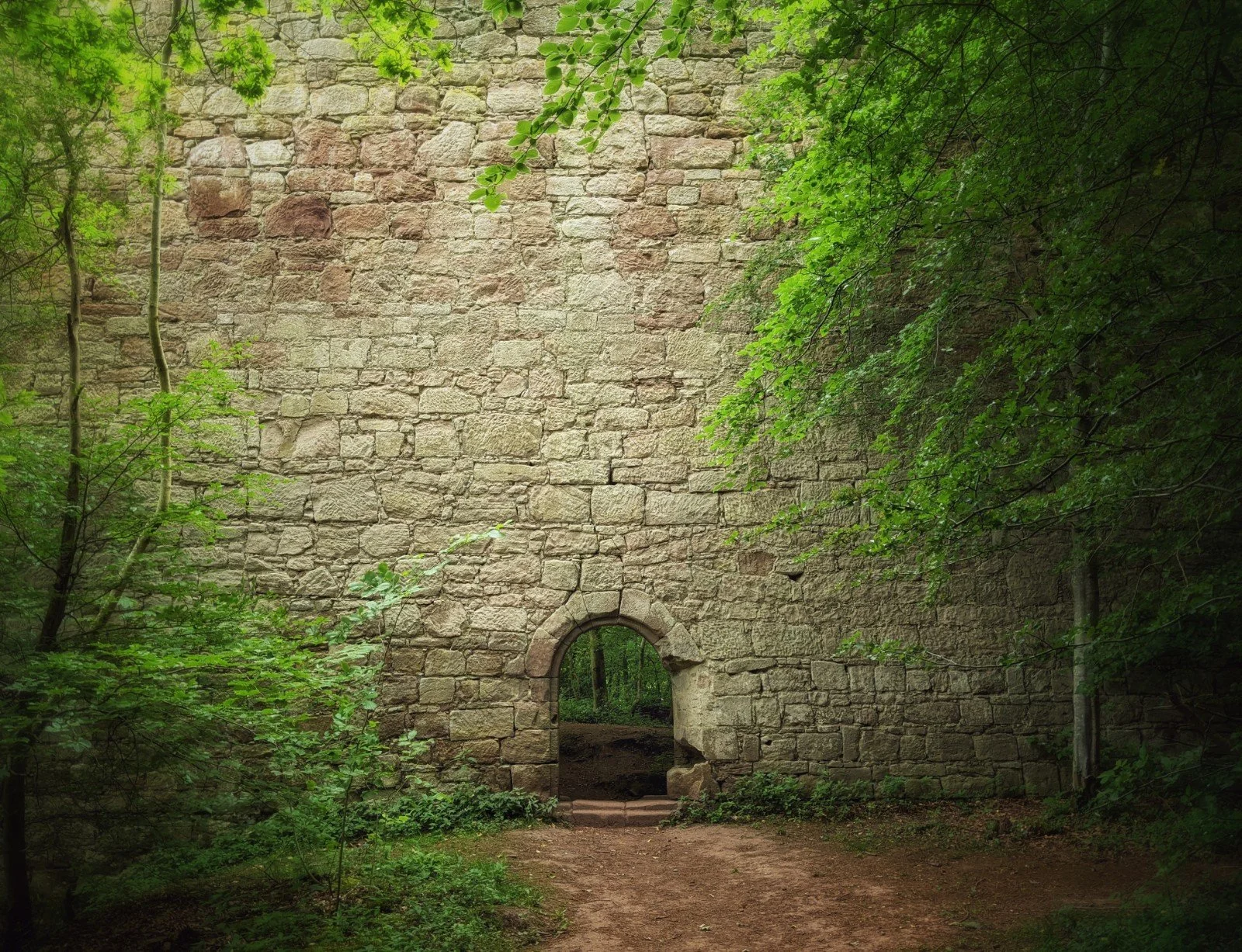 A mysterious, almost haunted-feeling red sandstone wall with no top or edges in frame and a single doorway through it, flanked by luch green foliage. Yester Castle, East Lothian.