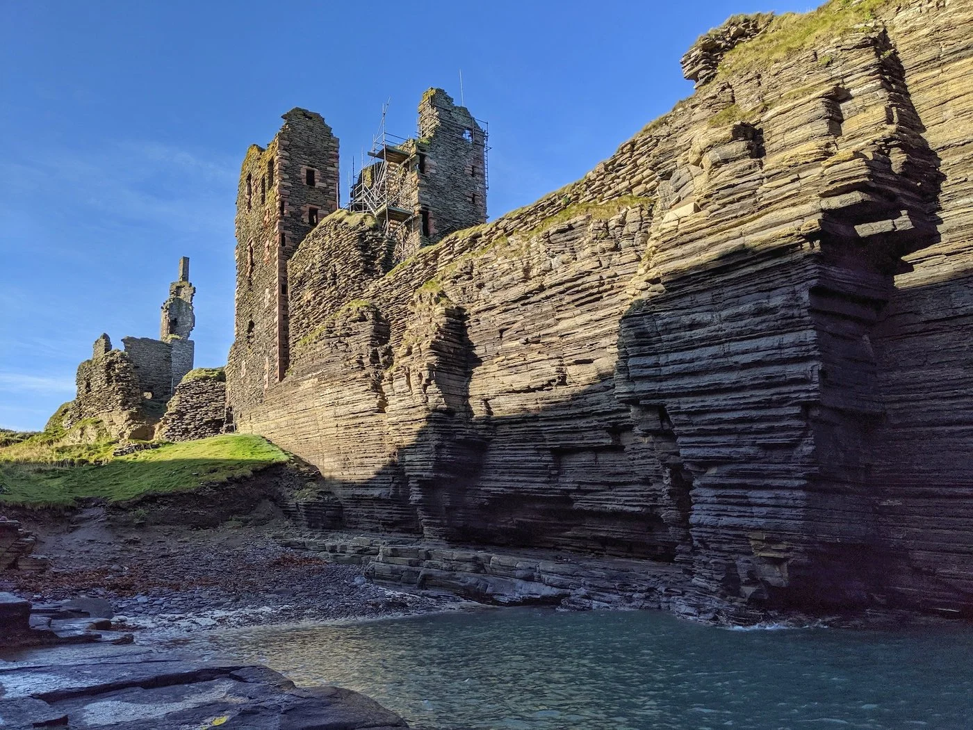 A strong but ruinous stone castle seems to grow out of rippling coastal cliffs. An inlet of water laps the cliffs, with blue skies above.