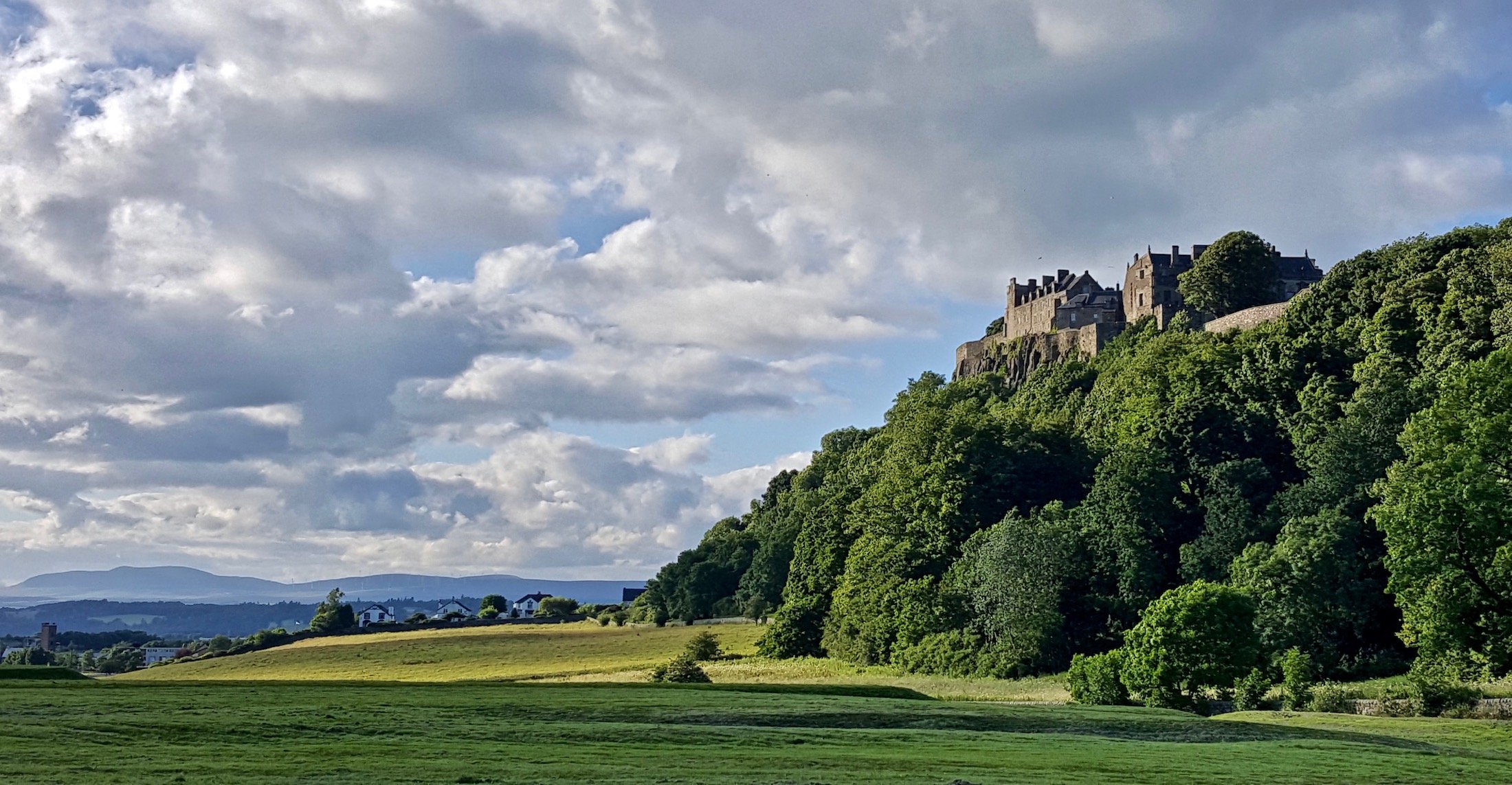 Stirling Castle atop a tall crag covered in lush green foliage, overlooking green fields and distant hills with cloudy blue skies.