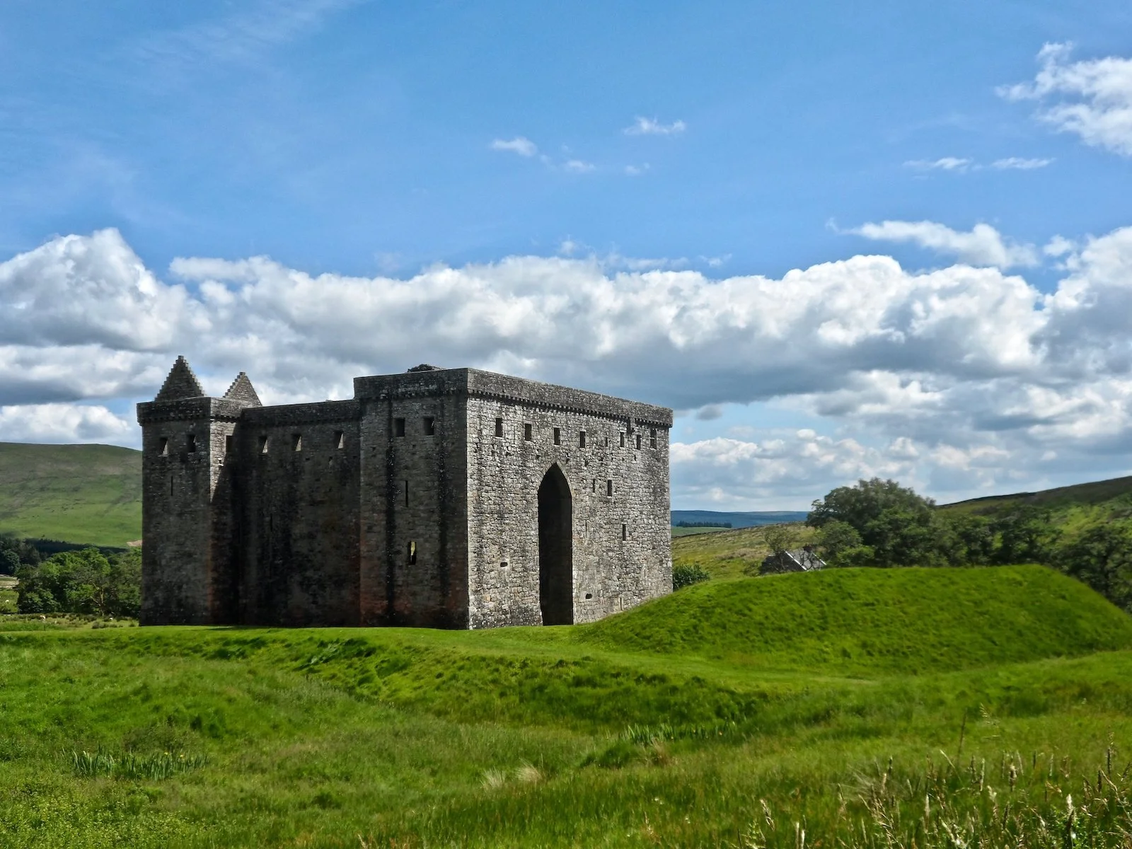 A formidable, brooding, squat square castle with a giant archway on one side amid a hilly, grassy field. Hermitage Castle, Scottish Borders.