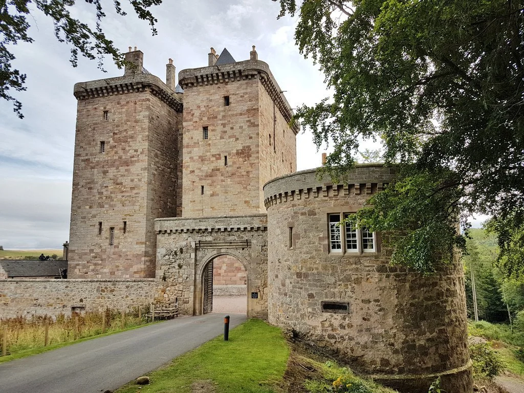 A narrow paved road leads through the gatehouse of Borthwick Castle, Midlothian. A short round tower flasnks the gate, witht he castle itself appearing like two narrow towers fitted together.