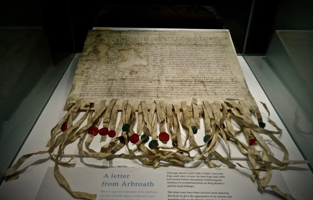 A frayed medieval document with tassels and wax seals appended to it, and black Latin script in dense rows across it. The Declaration of Arbroath at Arbroath Abbey.