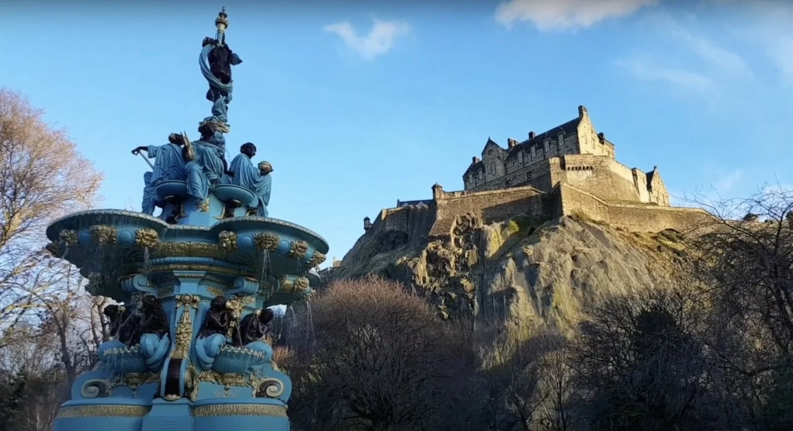 View from the vivid turquoise and gold Ross Fountain in Princes Street Gardens up to Edinburgh Castle on its crag, the castle walls illuminated by soft evening light.