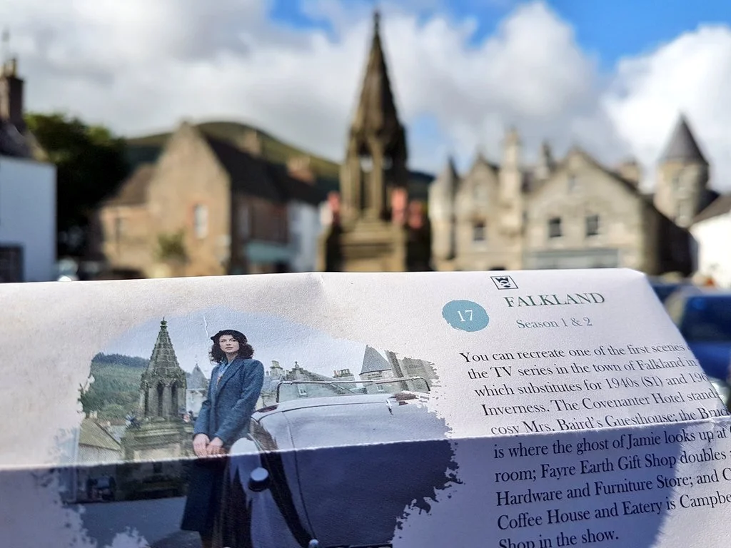 A paper guide to Falkland held up to the camera, depicting Claire from Outlander standing in the village, with the exact same scene Claire is in comprising the slightly blurred background.