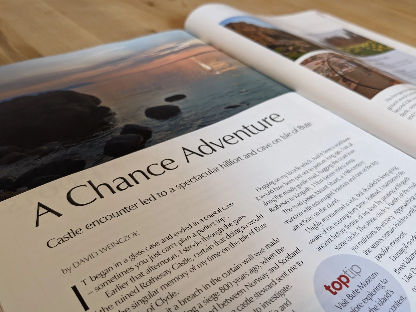 Open page of a magazine, The Scots Magazine, with the text blurred. The title reads, 'A Chance Adventure', backed by an image of a sunset over still waters on Bute.