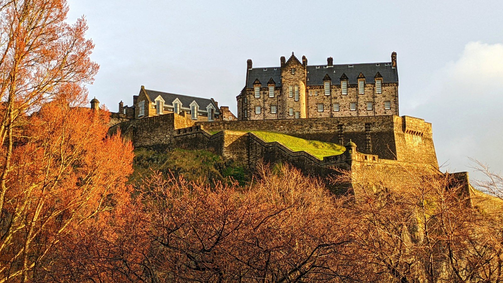Closeup up a section of Edinburgh Castle, vividly illuminating in orange-gold light with the red leaves of trees below forming a frame aroud the bottom and sides of the image.