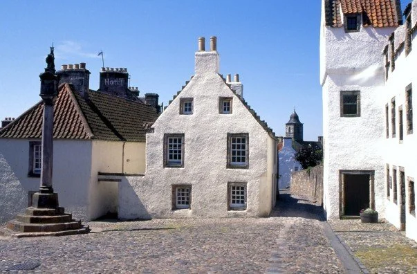 In the village of Culross, Fife, 17th century buildings with white walls and red tiled roofs surround a cobblestone courtyard with a stone cross on its left side.