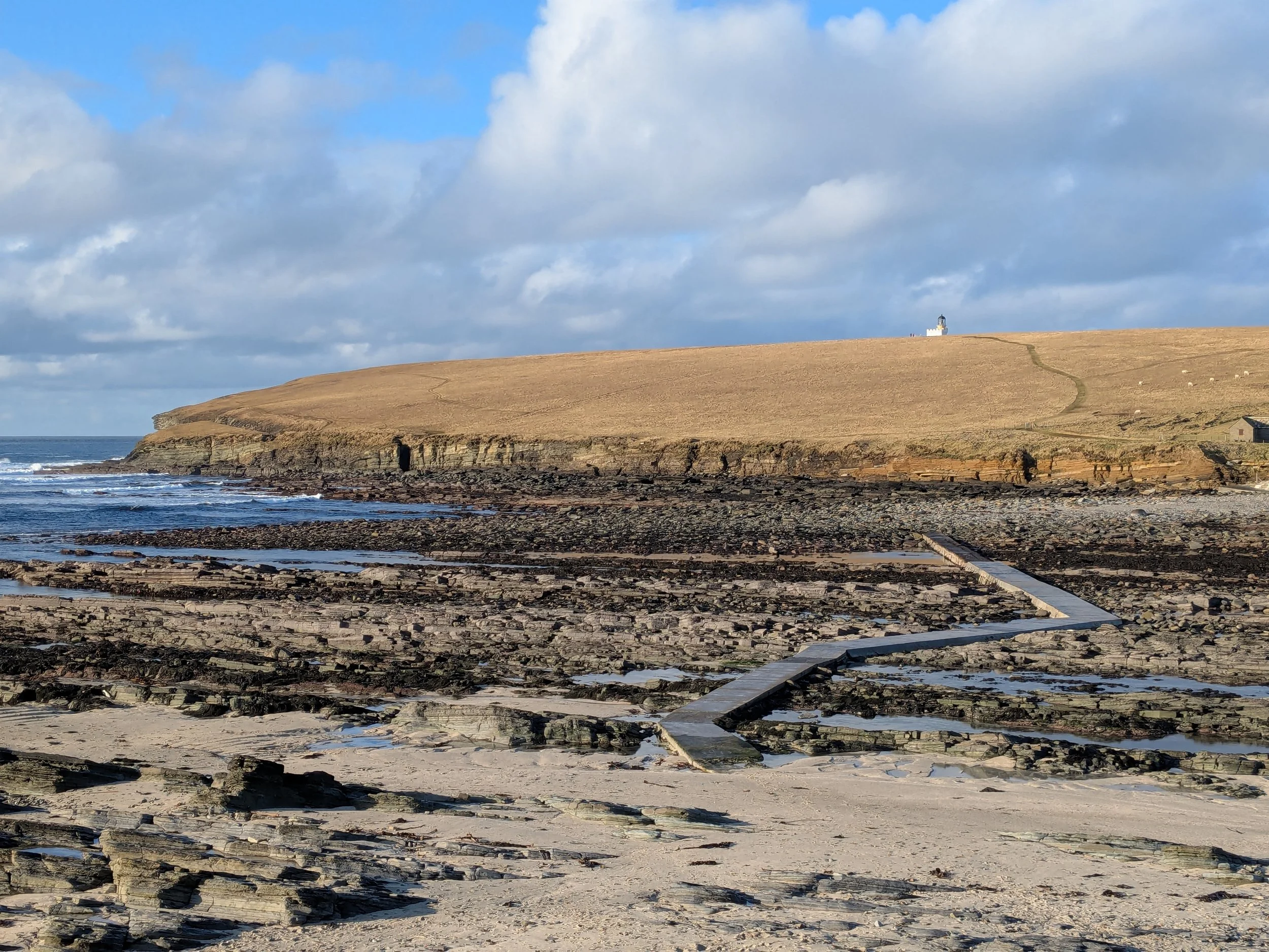 A low stone causeway extends across a rock-strewn sea bed exposed at low tide towards a grassy island with a lighthouse.