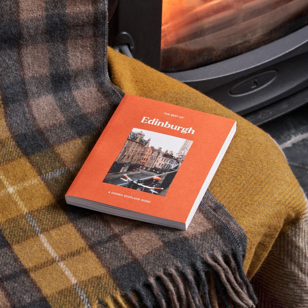 A book with an orange covere titled 'The Best of Edinburgh' rests at an angle on a chair covered with a grey and yellow tartan blanket.