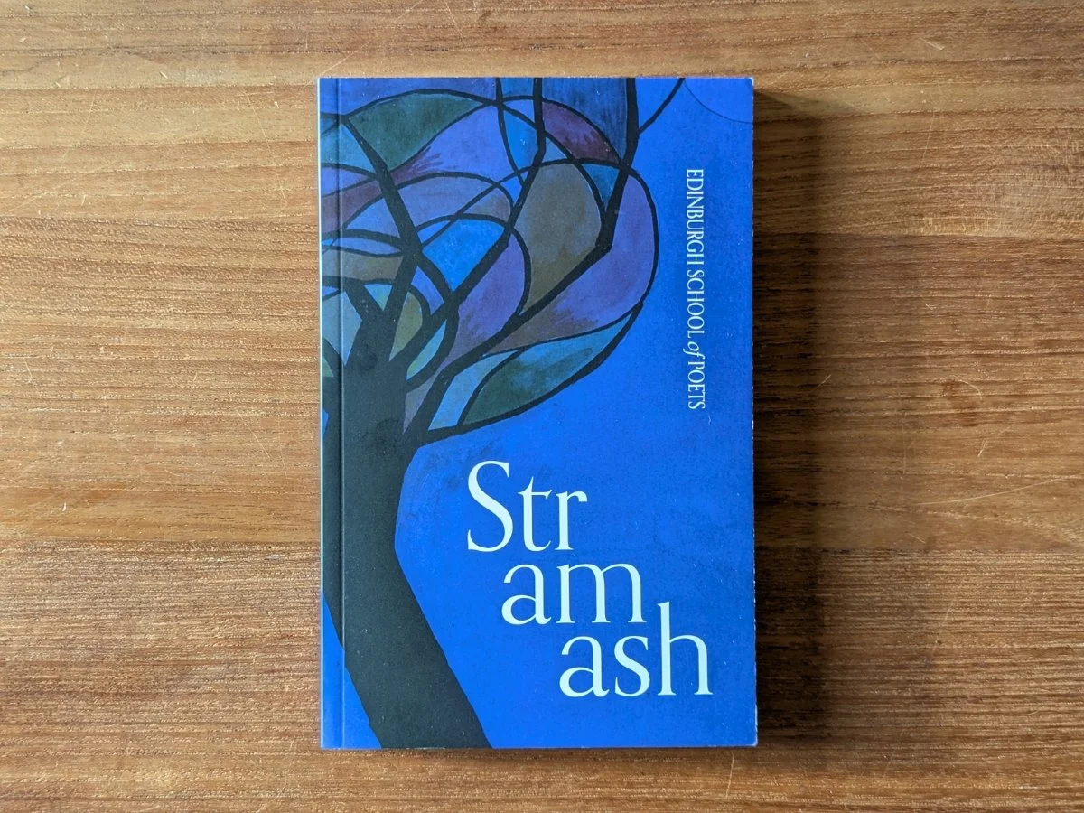 A blue book sits facing cover-up on a brown wooden surface. The book's title is 'Stramash', and a stained glass-style tree graces the cover.