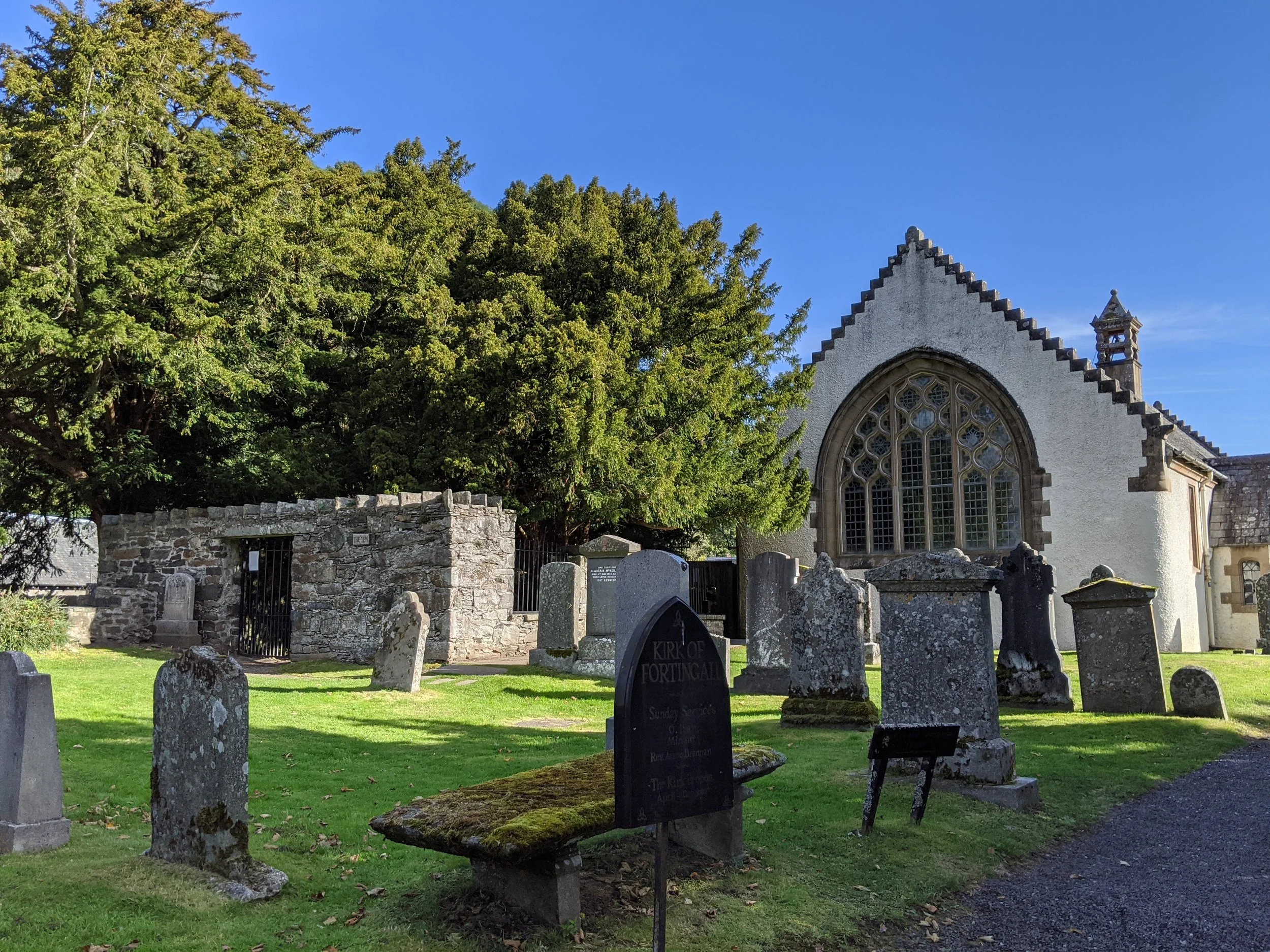 A small white church and graveyard around it with a stone enclosure containing a lush yew tree. Blue skies above and midday light on the church.