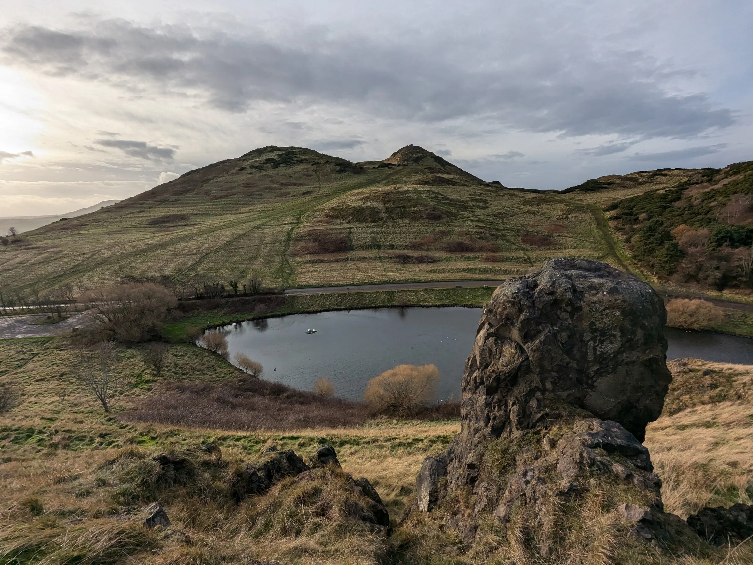 A view of the summit of Arthur's Seat with a small loch and natural stone pillar in the foreground.