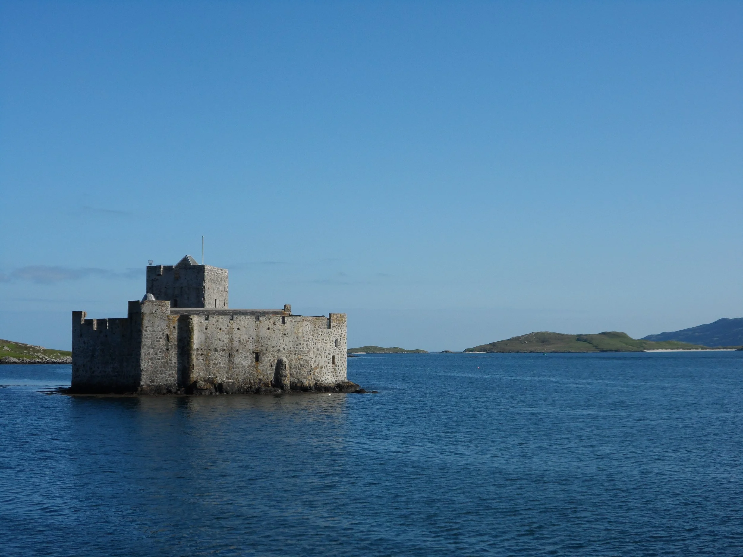 A small grey stone castle stands upon a rocky islet barely bigger than the castle itself, surrounded by blue water with several peninsulas in the distance.