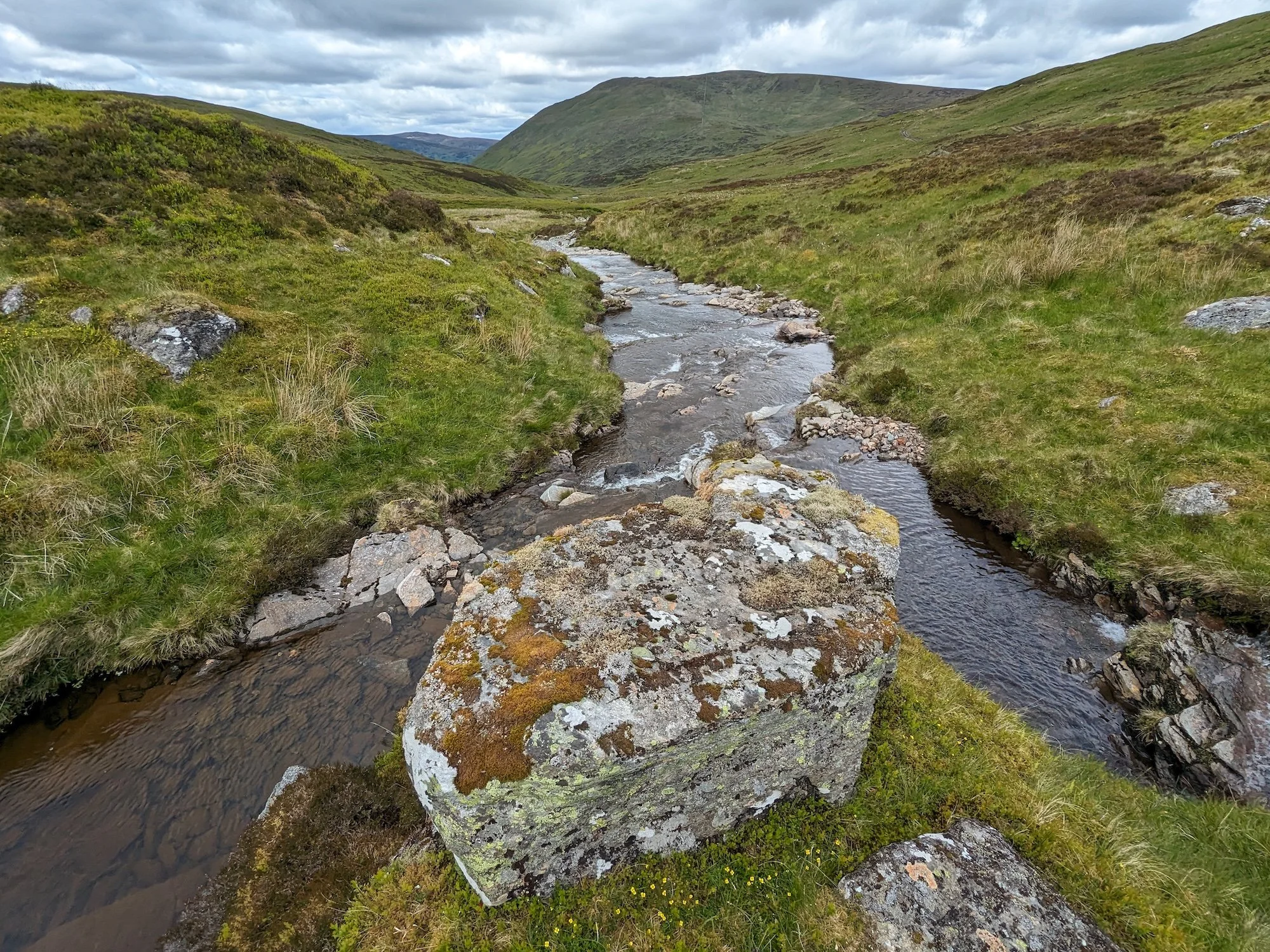 A fork in a small river viewed from a raised rocky knoll in the middle of the water. Upland hills extend into the distance with green grass and brown bracken.