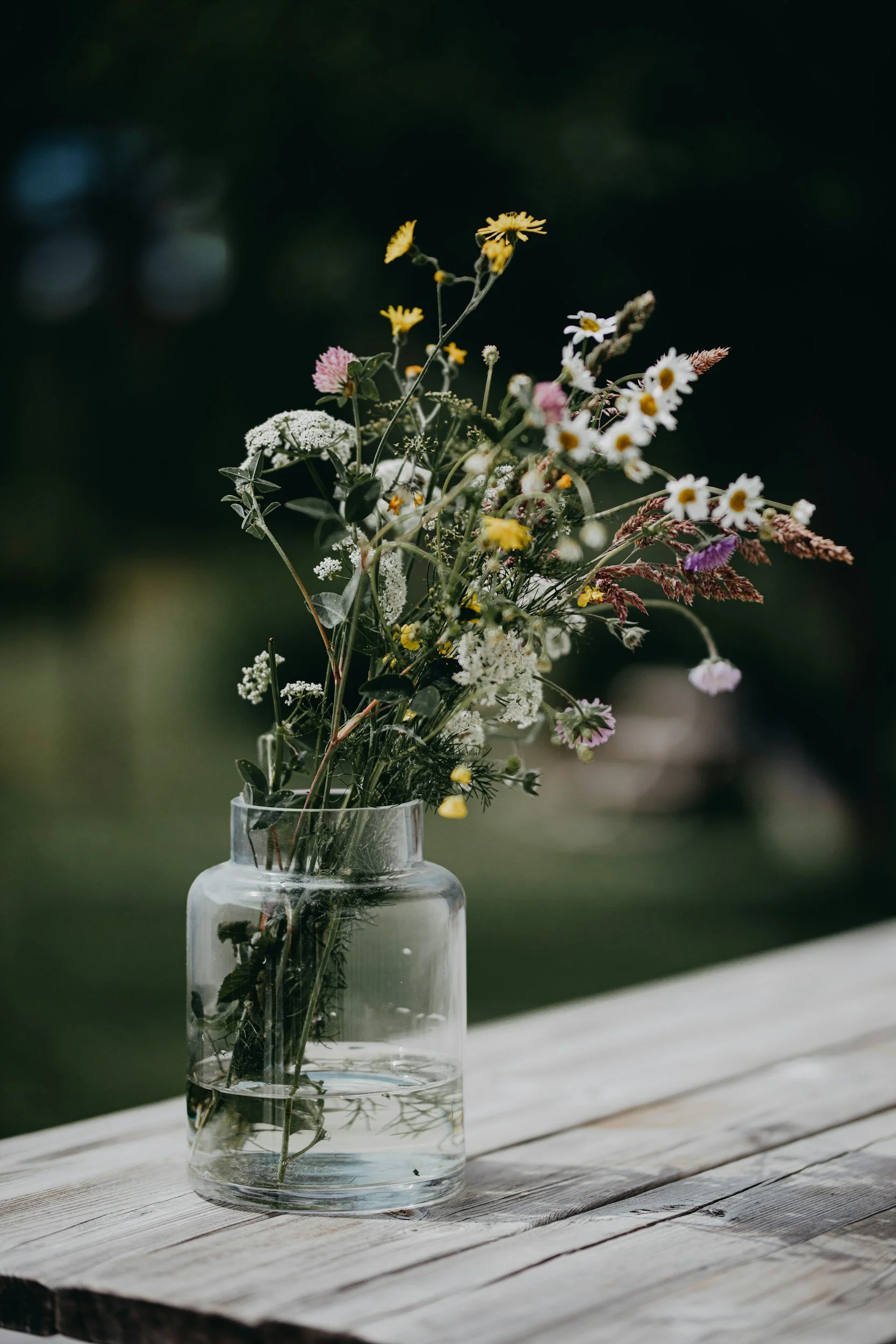 A clear glass vase with a bouquet of mixed wildflowers on a weathered white wooden surface outdoors with a blurred dark green background.
