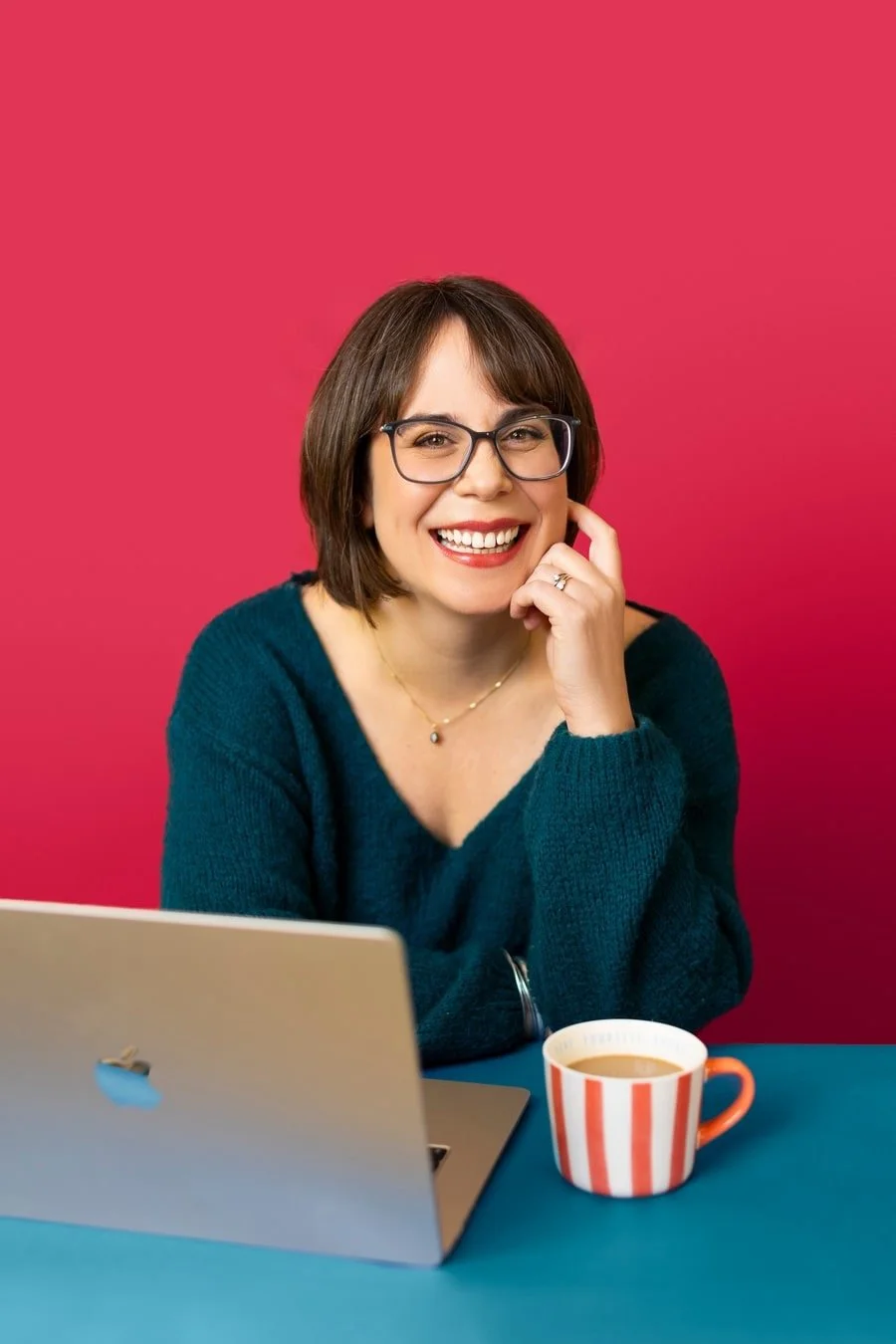 A woman with short brown hair, wearing glasses and a teal sweater, sitting at a desk with a laptop and a striped mug, smiling at the camera against a pink background.