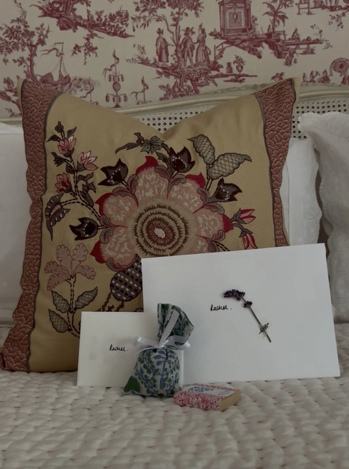 Decorative pillows on a bed with printed fabric pattern, and gift boxes wrapped with ribbons, placed in front of the pillows. The background shows a fabric wall hanging with pastoral scenes. There are greeting cards with the name "Rachel" on them and a small wrapped item with a ribbon.