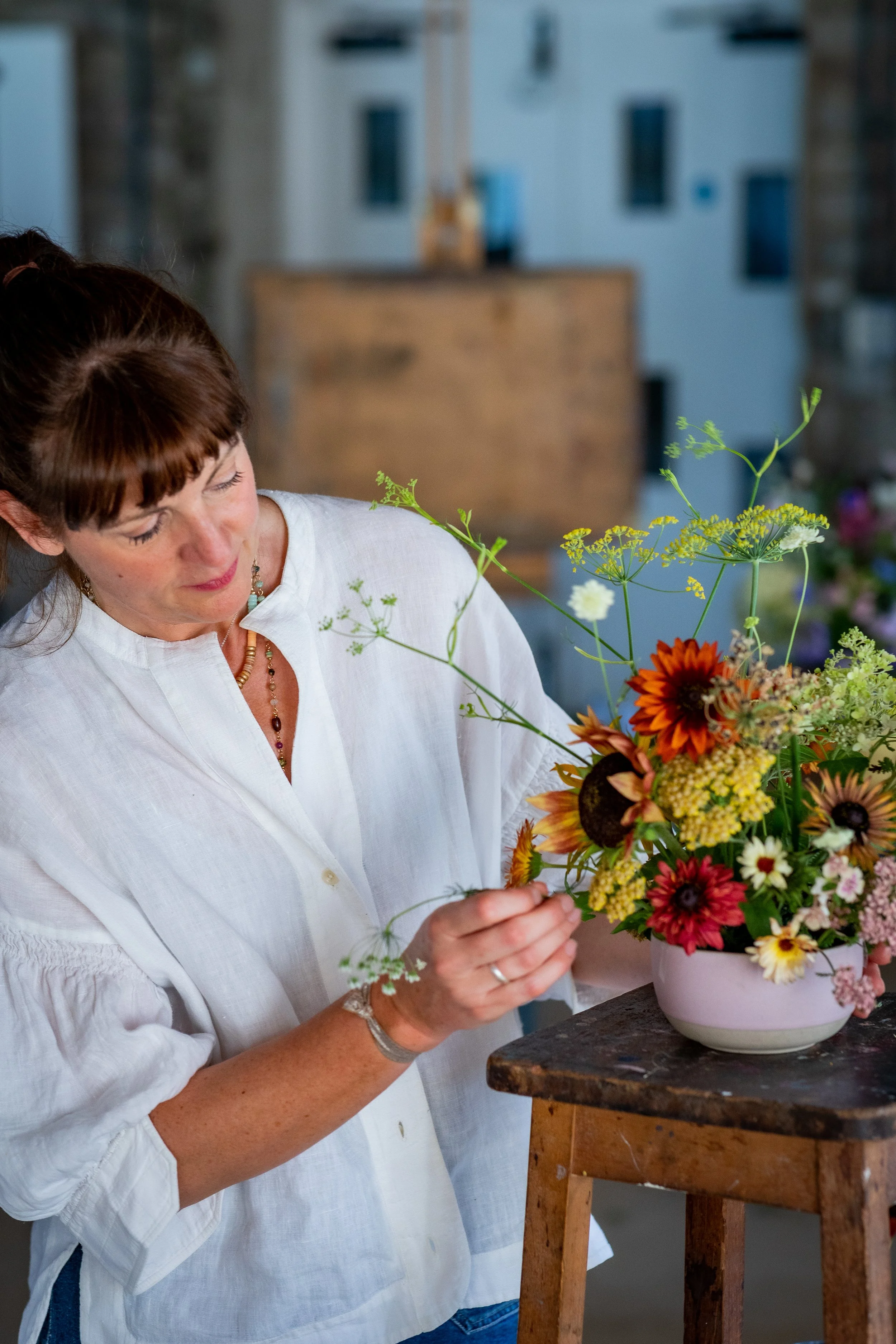 A woman with brown hair and a white blouse arranging a colorful flower bouquet in a pink vase on a wooden stool.