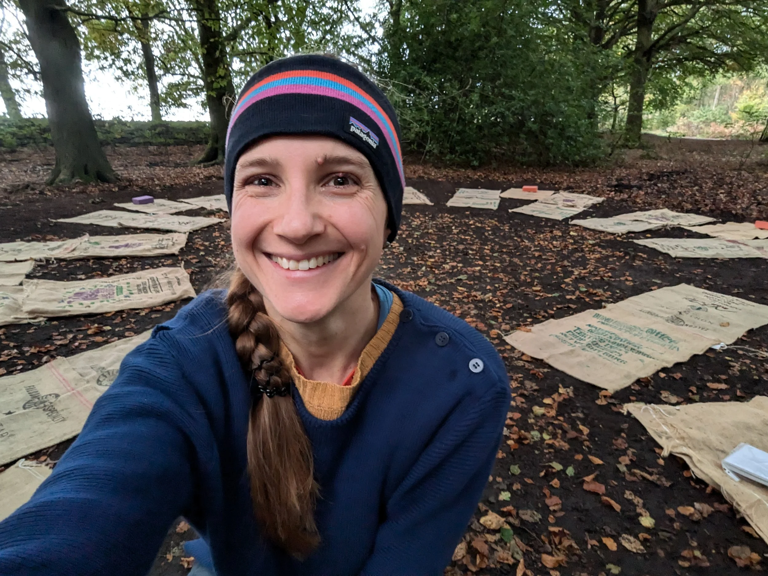 A person wearing a blue sweater and a hat with colorful stripes smiles in a forest setting. The ground is covered with fallen leaves and several burlap sacks are laid out in a circular arrangement around them. Trees with green foliage are in the background.