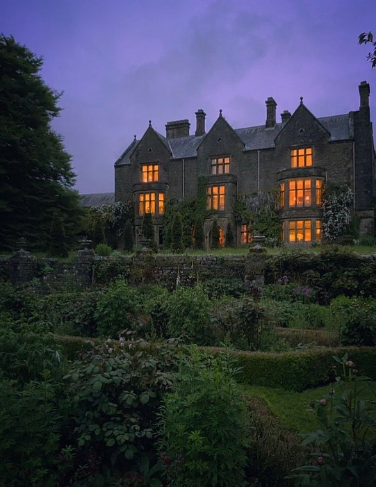 A large, dark stone mansion with multiple towers and glowing windows, surrounded by lush greenery and a garden, under a purple evening sky.