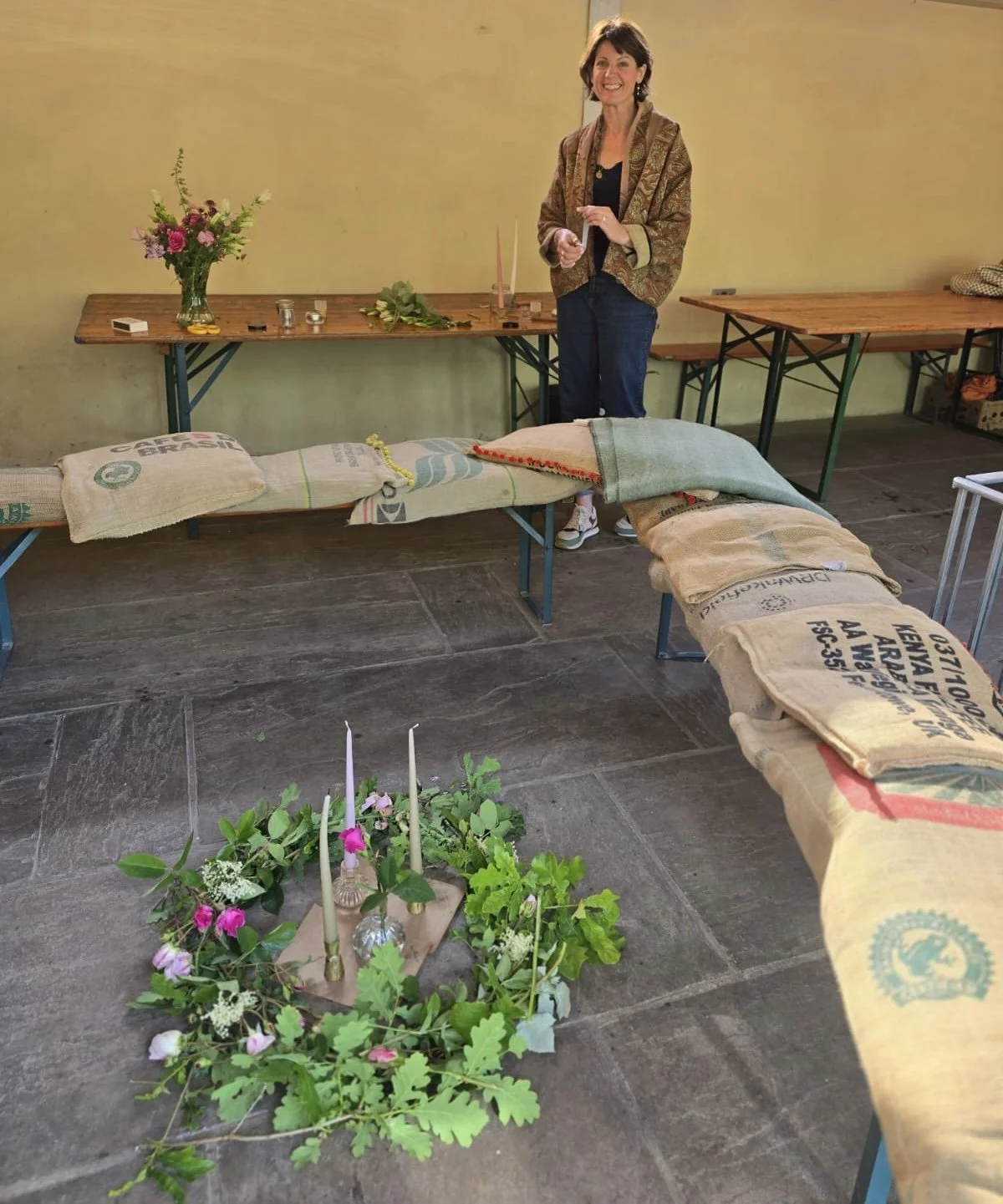 A woman standing behind a table arranged with candles, flowers, and other items, with a row of burlap sacks and a floral arrangement with candles on the floor in front.
