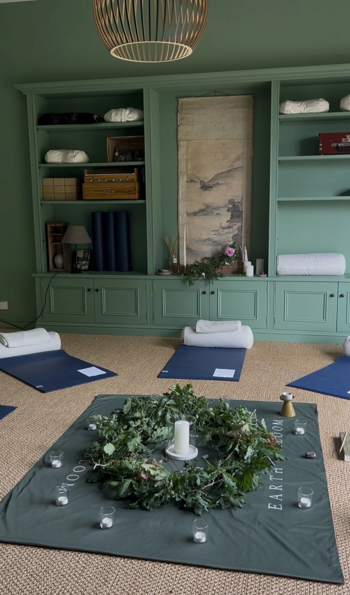 A room set up for a yoga or meditation session with a dark green mat on the floor, surrounded by small candles, a large white candle in the center, and green plants. In the background, there are shelves with folded blankets and other items, a hanging scroll with a landscape painting, and a beige carpet.