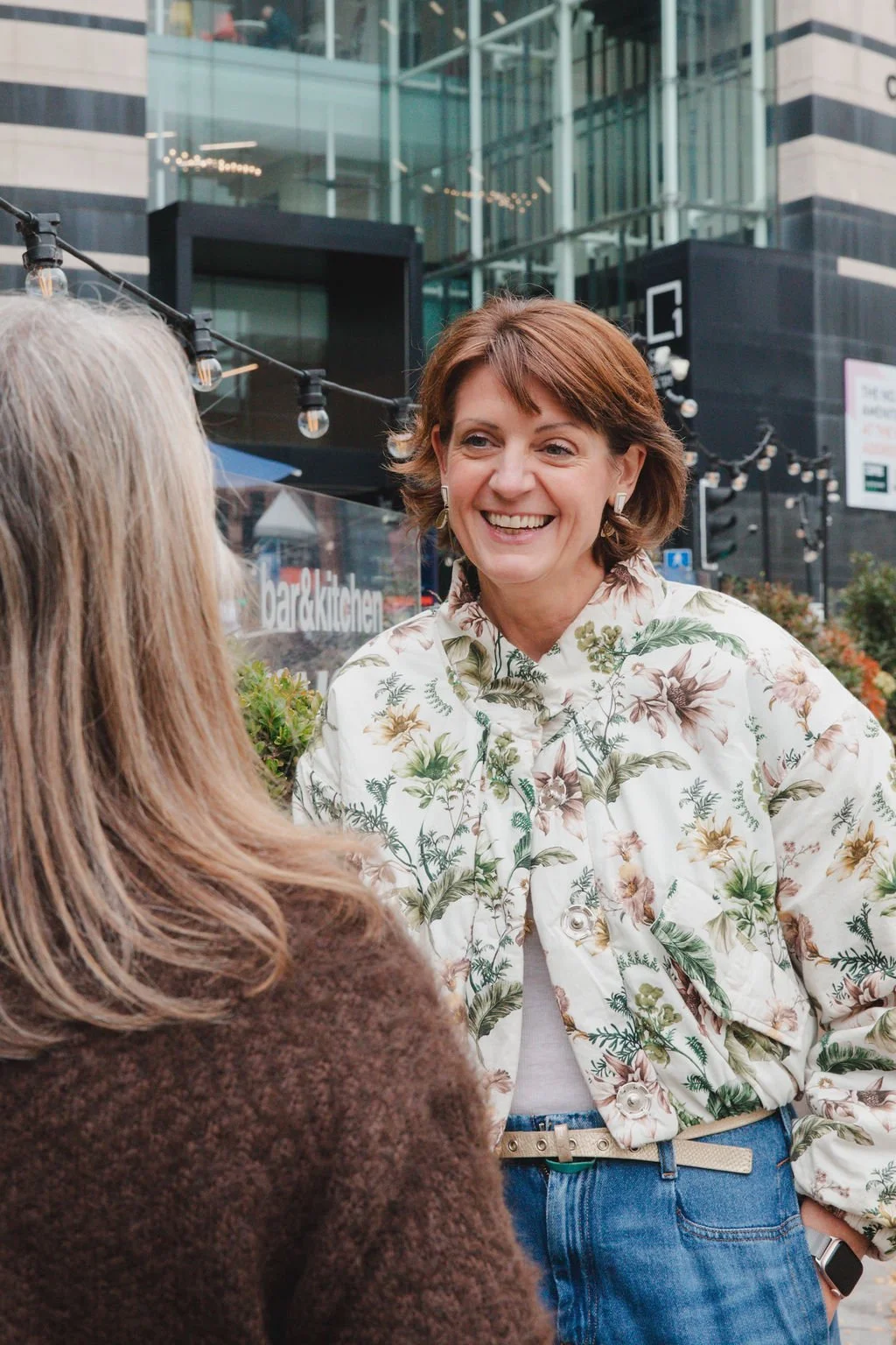 Two women engaged in a conversation outdoors in an urban setting, one smiling, wearing a floral patterned shirt and blue jeans, the other with long light brown hair, wearing a brown textured top. Modern buildings and hanging string lights are visible in the background.